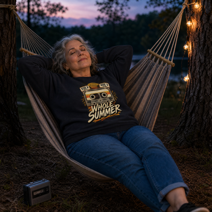 A woman relaxes in a wooden string hammock outdoors at dusk, wearing a black long-sleeve graphic sweatshirt with a retro cassette tape design and the words “Whole Summer” in yellow and beige. Trees, string lights, and a small cassette tape on the ground are visible.