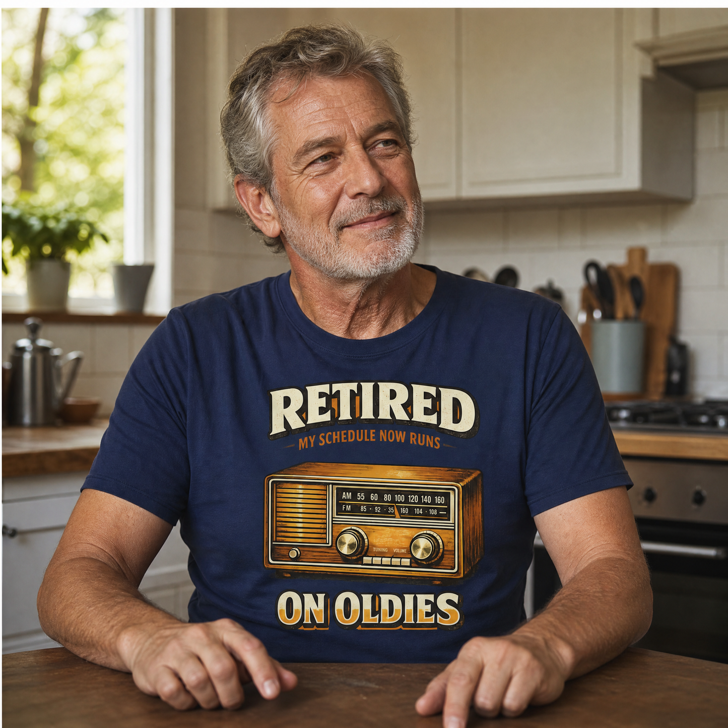 A man sitting at a kitchen table wearing a navy blue graphic t-shirt with a retro radio design and the text 'RETIRED MY SCHEDULE NOW RUNS ON OLDIES'.