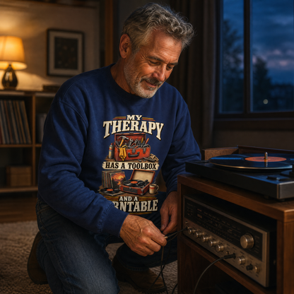 A man kneels on the floor in a cozy, dimly lit room, connecting an audio cable. He is wearing a navy blue crewneck sweatshirt with a graphic print that reads 'MY THERAPY HAS A TOOLBOX '.
