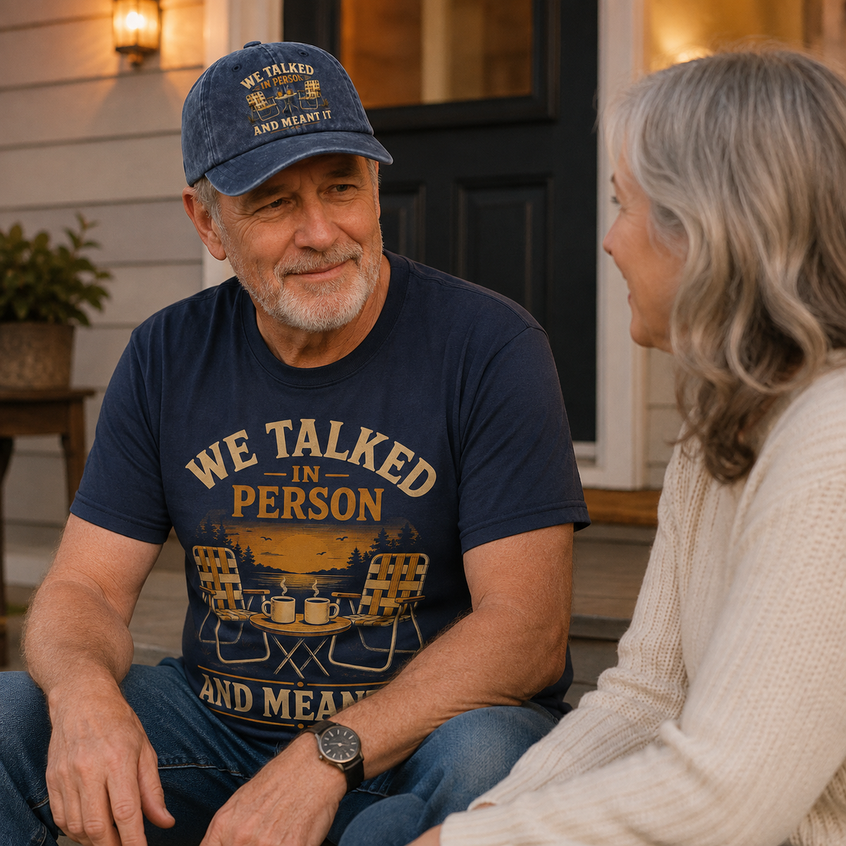 An older man wearing a navy blue graphic t-shirt and matching baseball cap sits on a porch talking to a woman. The t-shirt features a vintage-style graphic with camping chairs and the text 'WE TALKED IN PERSON AND MEANT IT'.