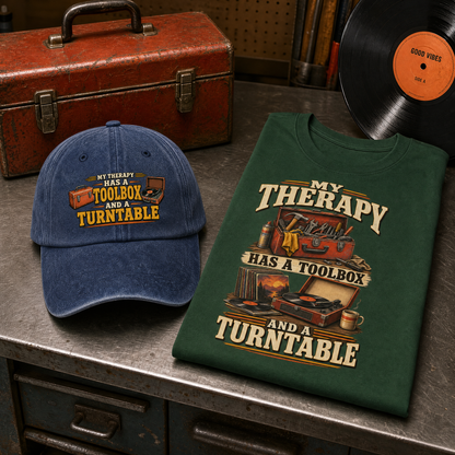 A folded dark green graphic T-shirt and a blue baseball cap displayed on a metal workbench, with a vintage red toolbox and a vinyl record in the background. The cap and shirt both feature the phrase 'MY THERAPY HAS A TOOLBOX AND A TURNTABLE' with illustrated tools and a light bulb.