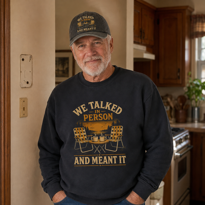 An older man standing in a kitchen wearing a dark charcoal crewneck sweatshirt with a yellow graphic that reads 'WE TALKED IN PERSON AND MEANT IT' with camping chairs and a campfire, along with a matching baseball cap.