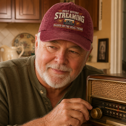 A smiling older man with a white beard wears a maroon baseball cap with the text 'BUILT BEFORE STREAMING RAISED ON THE REAL THING' and holds a vintage-style radio in a warm indoor setting.
