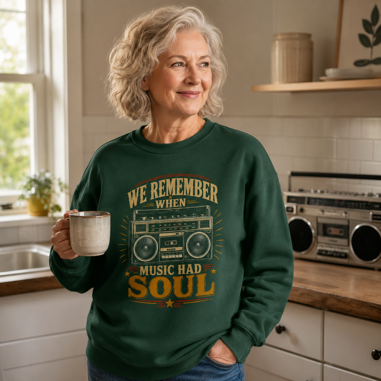 An older woman with short gray hair stands in a bright kitchen holding a ceramic mug. She is wearing a dark green crewneck sweatshirt with a vintage-style graphic that reads 'WE REMEMBER WHEN MUSIC HAD SOUL' around a retro boombox illustration.