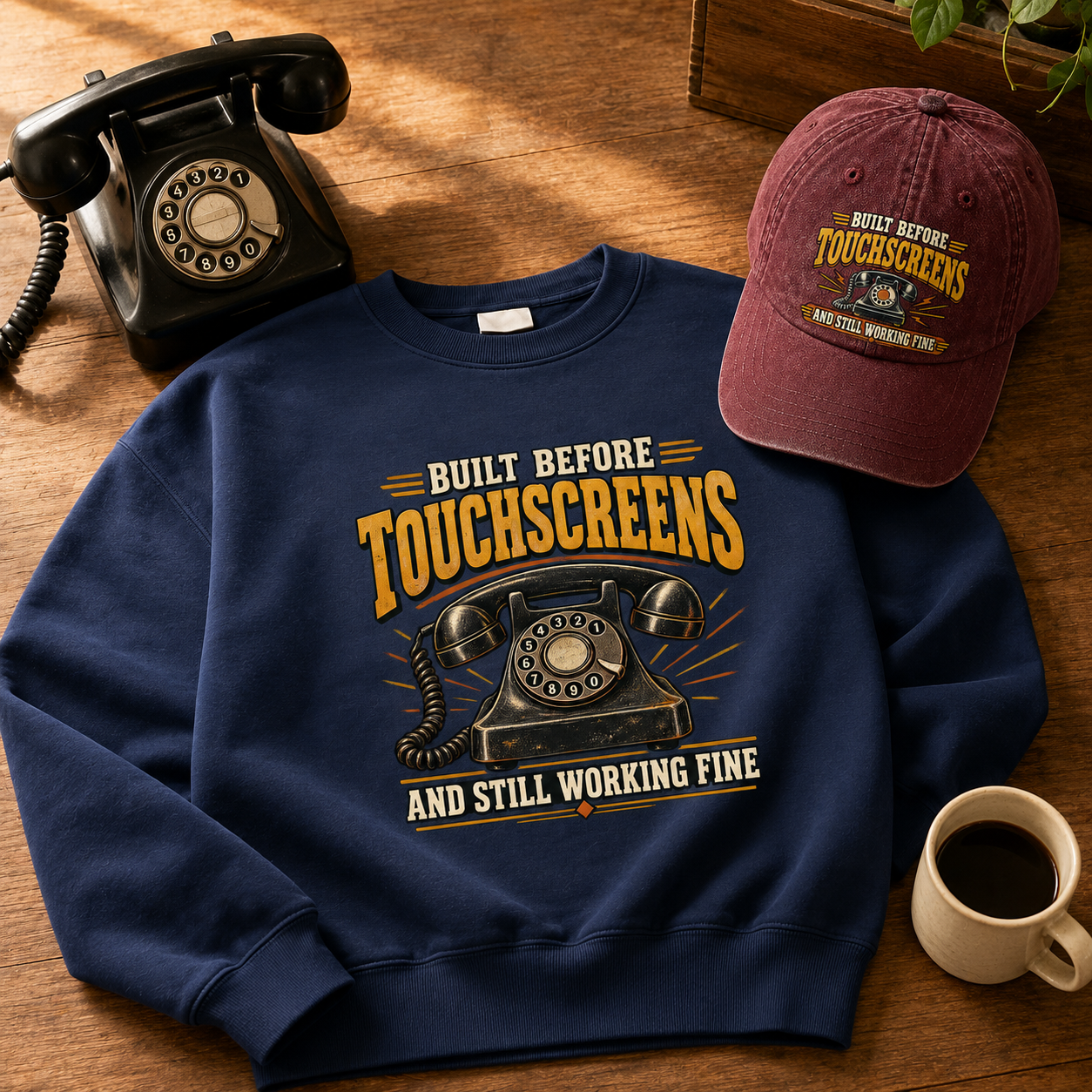 A navy blue crewneck sweatshirt with a retro graphic reading 'Built Before Touchscreens And Still Working Fine' is laid flat on a wooden surface next to a black rotary phone, a maroon baseball cap with matching text, and a coffee cup.