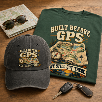 A dark forest green graphic T-shirt and a dark charcoal gray baseball cap, both printed with the phrase 'BUILT BEFORE GPS WE STILL GOT THERE' and a map/compass design, arranged on a wooden tabletop with sunglasses, a map, and car keys as props.