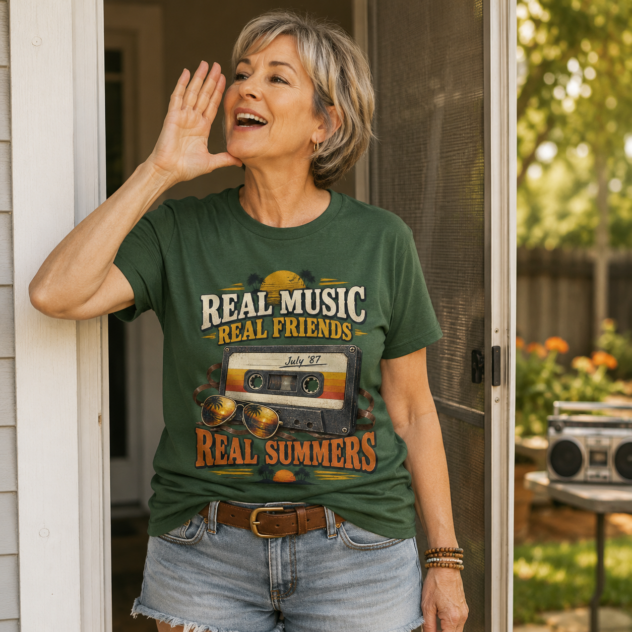 An older woman stands in a doorway wearing a dark green graphic T-shirt with the text 'REAL MUSIC REAL FRIENDS' and 'REAL SUMMERS' around a cassette tape illustration. She is also wearing light-wash denim shorts, and a sunny backyard is visible behind her.