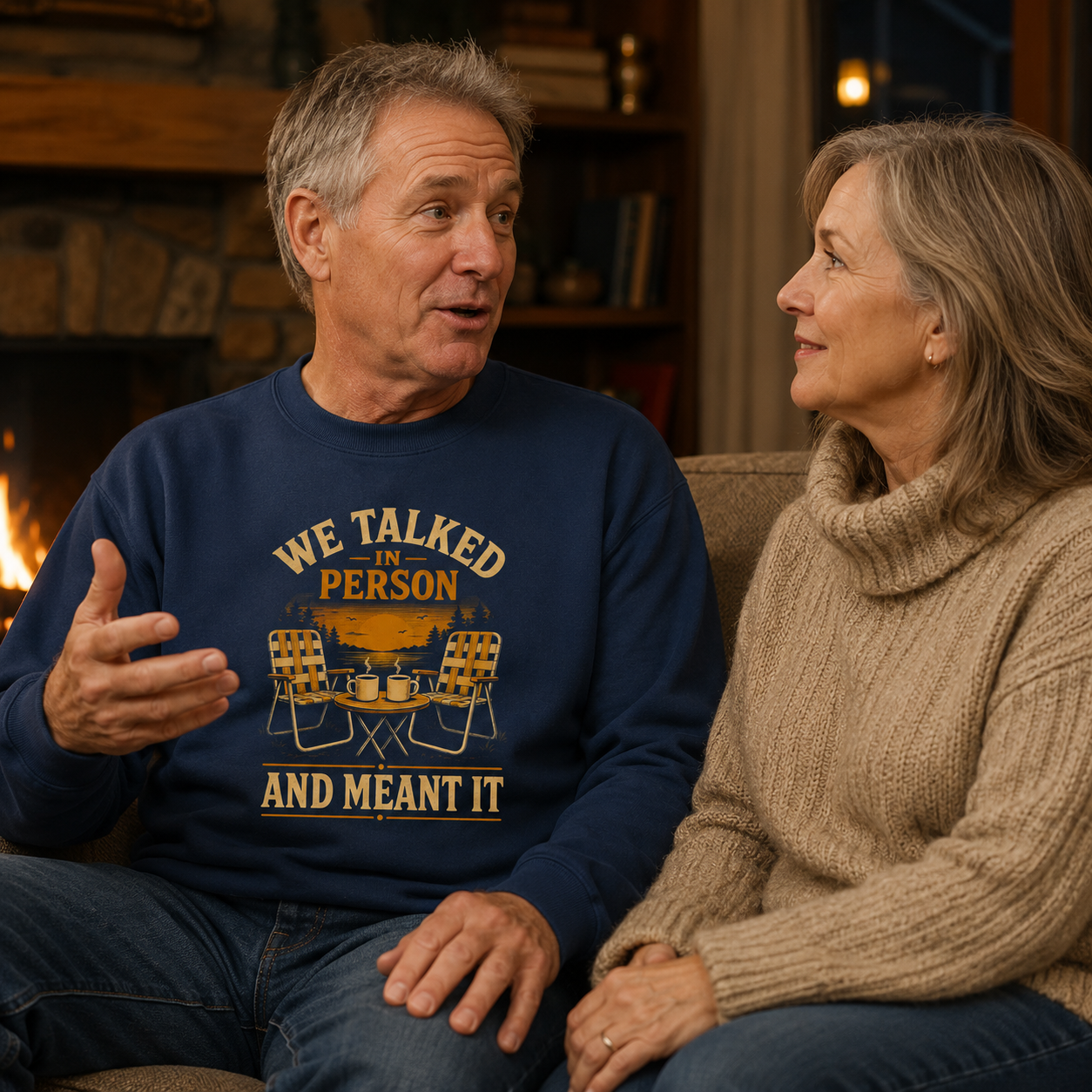 A man and a woman sitting together in a cozy living room setting, with the man wearing a navy blue crewneck sweatshirt that features a yellow and white graphic reading 'WE TALKED IN PERSON AND MEANT IT' with camping chairs and a campfire design.