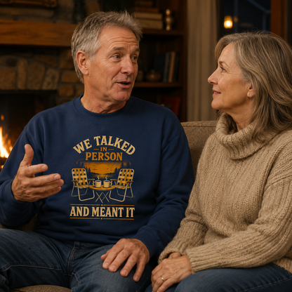 A man and a woman sitting together in a cozy living room setting, with the man wearing a navy blue crewneck sweatshirt that features a yellow and white graphic reading 'WE TALKED IN PERSON AND MEANT IT' with camping chairs and a campfire design.