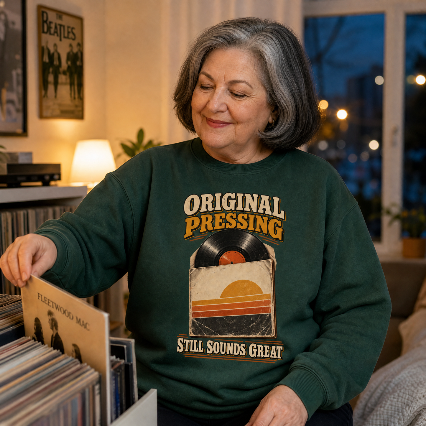 An older woman with gray hair is seen in a cozy room, looking down at a vinyl record in a record rack. She is wearing a dark green crewneck sweatshirt with a retro graphic that reads 'ORIGINAL PRESSING' and 'STILL SOUNDS GREAT' with a vinyl record and sunset design.