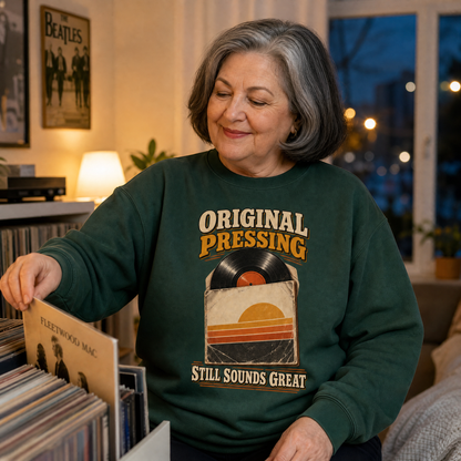 An older woman with gray hair is seen in a cozy room, looking down at a vinyl record in a record rack. She is wearing a dark green crewneck sweatshirt with a retro graphic that reads 'ORIGINAL PRESSING' and 'STILL SOUNDS GREAT' with a vinyl record and sunset design.
