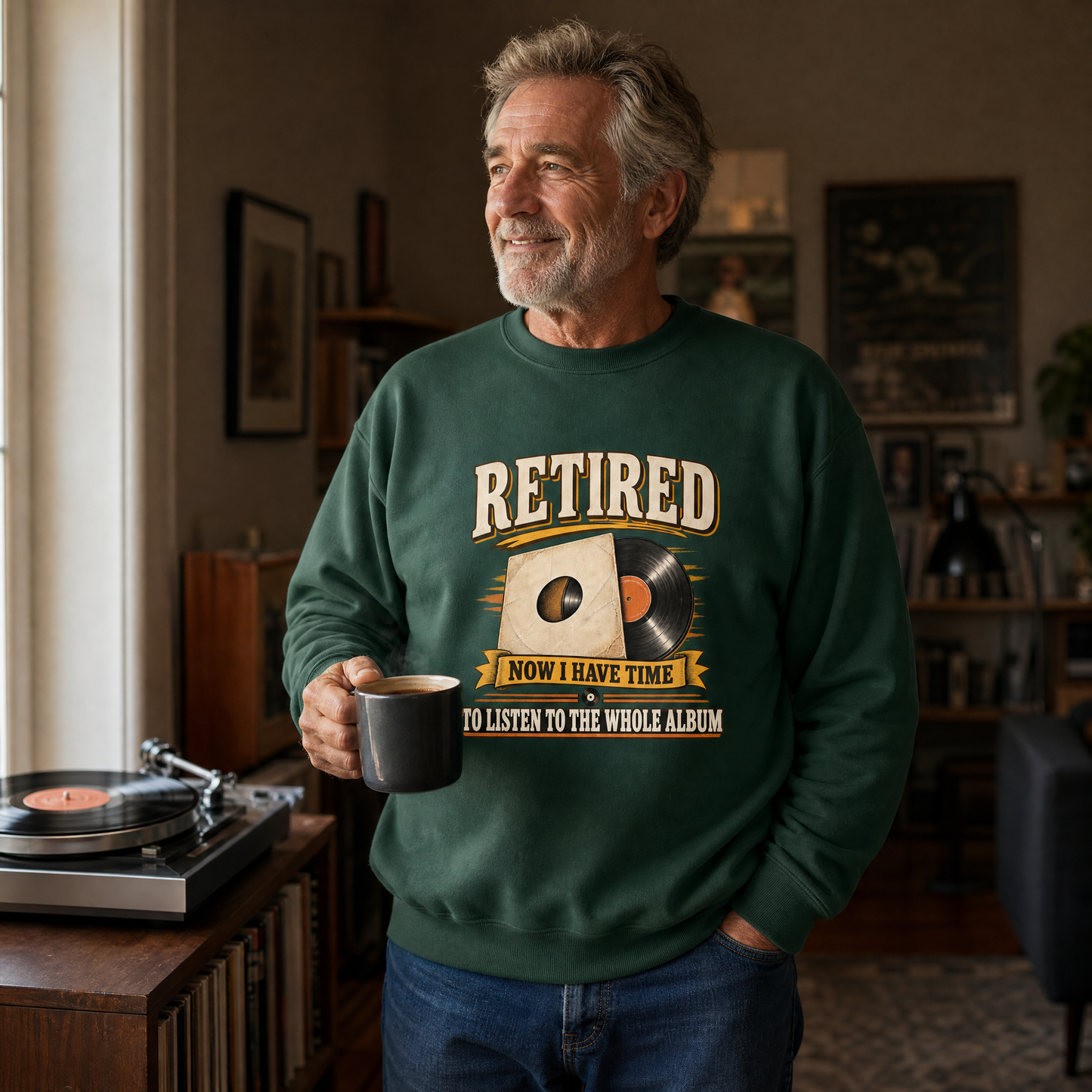An older man in a living room wearing a dark green crewneck sweatshirt with a retro graphic that reads 'RETIRED NOW I HAVE TIME TO LISTEN TO THE WHOLE ALBUM'. He is holding a black mug next to a turntable.
