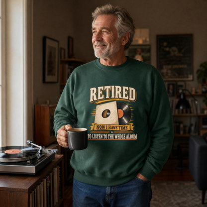 An older man in a living room wearing a dark green crewneck sweatshirt with a retro graphic that reads 'RETIRED NOW I HAVE TIME TO LISTEN TO THE WHOLE ALBUM'. He is holding a black mug next to a turntable.