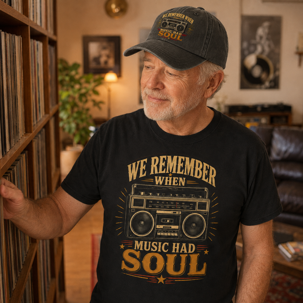 An older man wearing a black graphic t-shirt and a black baseball cap stands next to a wooden record shelf in a cozy living room. The t-shirt features a vintage-style graphic of a boombox with the text 'WE REMEMBER WHEN MUSIC HAD SOUL' in a warm beige and gold color.