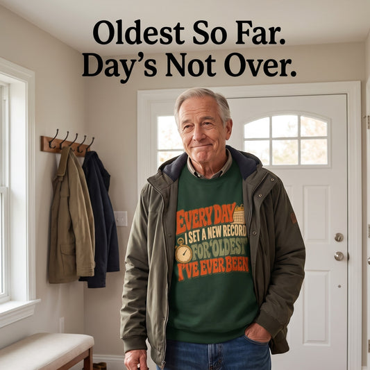 Senior man wearing green sweatshirt in entryway beneath Oldest So Far wall text