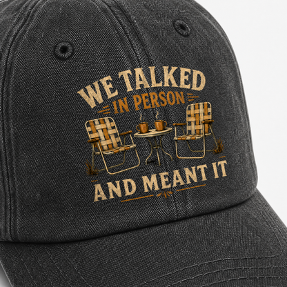 Black vintage washed cap with printed “WE TALKED IN PERSON” slogan and chairs, angled view.