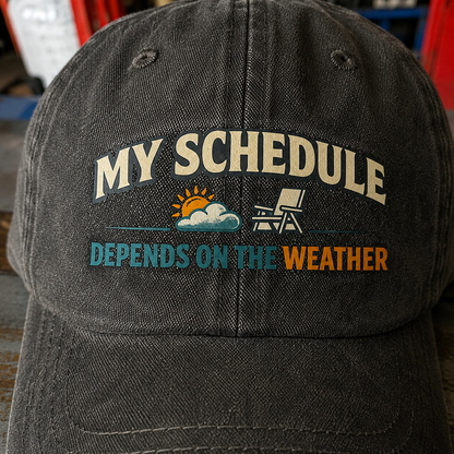 Black vintage washed cap with printed “My Schedule Depends On The Weather” slogan, front angle.