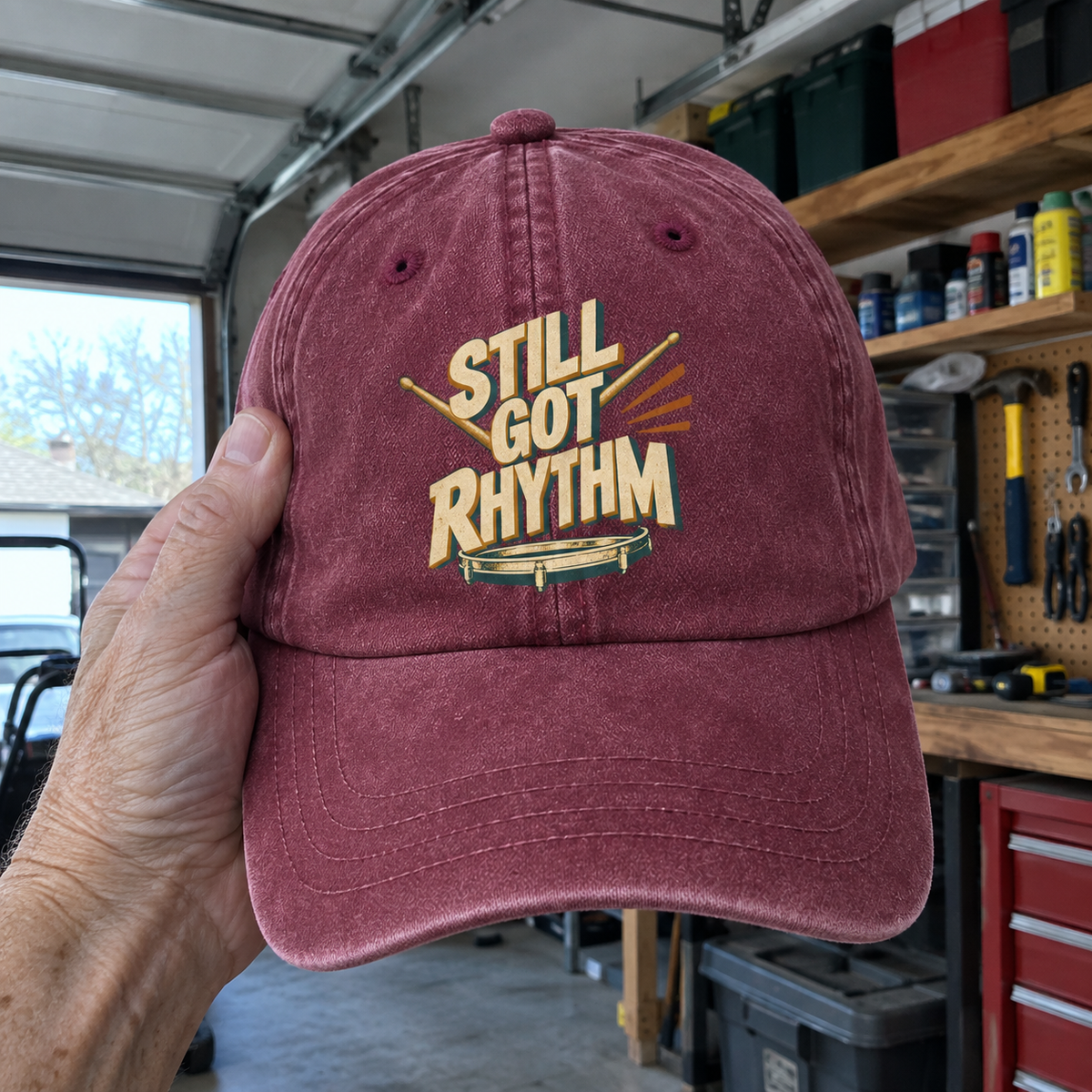 Burgundy vintage washed cap featuring printed Still Got Rhythm slogan, held by hand in garage