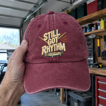 Burgundy vintage washed cap featuring printed Still Got Rhythm slogan, held by hand in garage