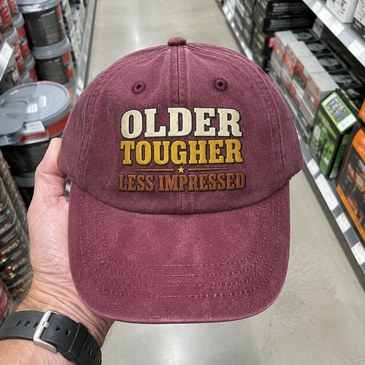 Burgundy vintage washed cap with printed Older Tougher Less Impressed slogan, handheld front view.