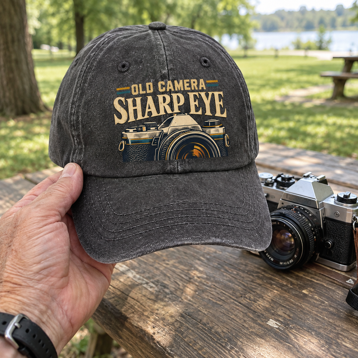 Vintage washed cap with printed Old Camera Sharp Eye slogan and camera design, handheld outdoors beside camera