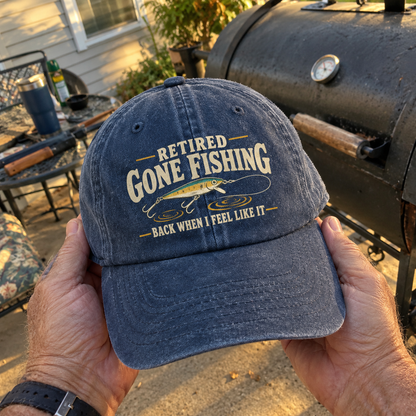 Blue vintage washed cap with printed Retired Gone Fishing slogan, handheld outdoors.