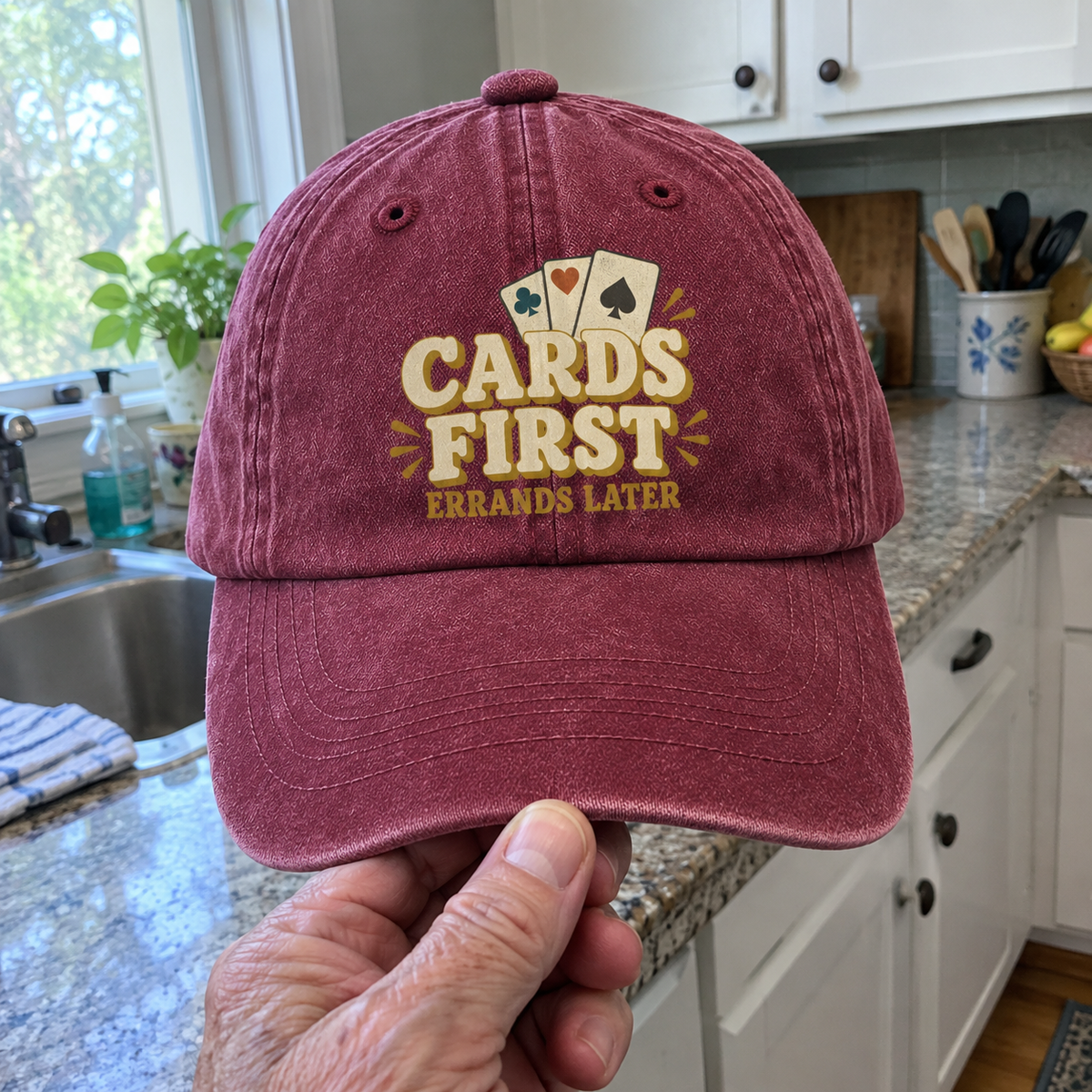 Burgundy vintage washed cap with printed “Cards First Errands Later” design, handheld front view