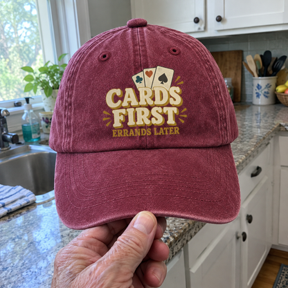 Burgundy vintage washed cap with printed “Cards First Errands Later” design, handheld front view
