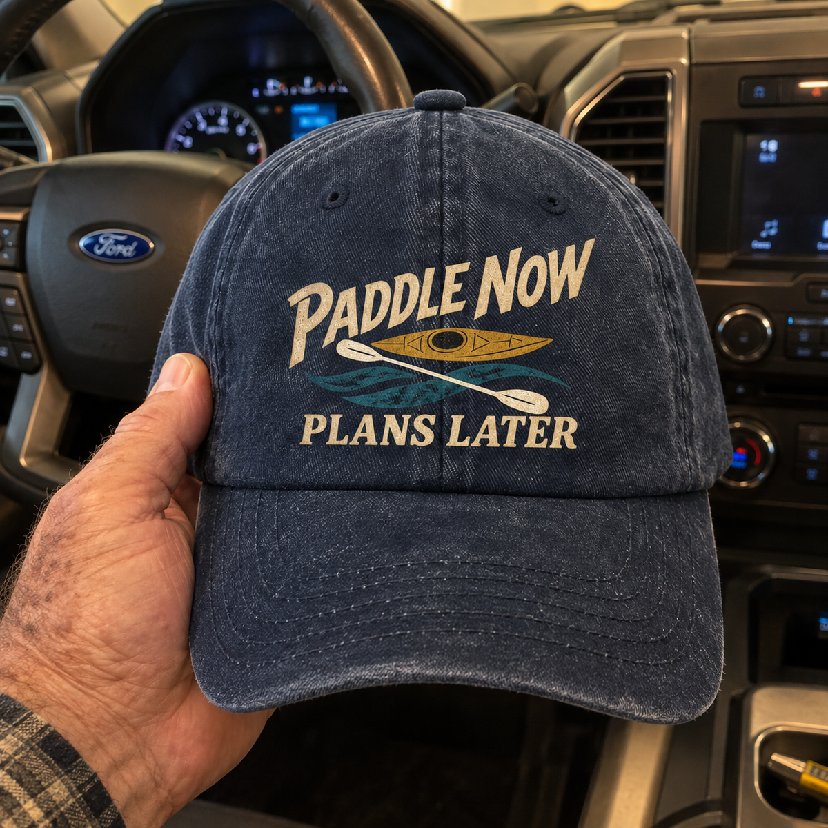 Navy vintage washed cap with Paddle Now Plans Later slogan, held inside a car