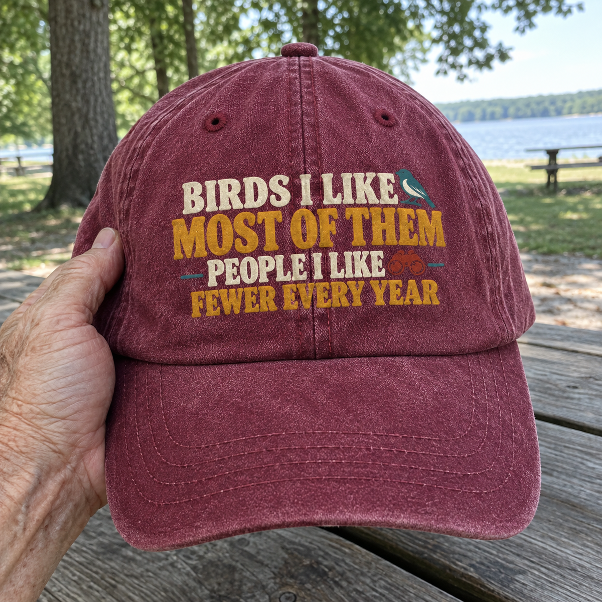 Burgundy vintage washed cap with printed bird slogan, handheld outdoors by a senior hand beside a lake