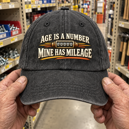 Charcoal vintage washed cap held by hands, printed Age Is A Number Mine Has Mileage.
