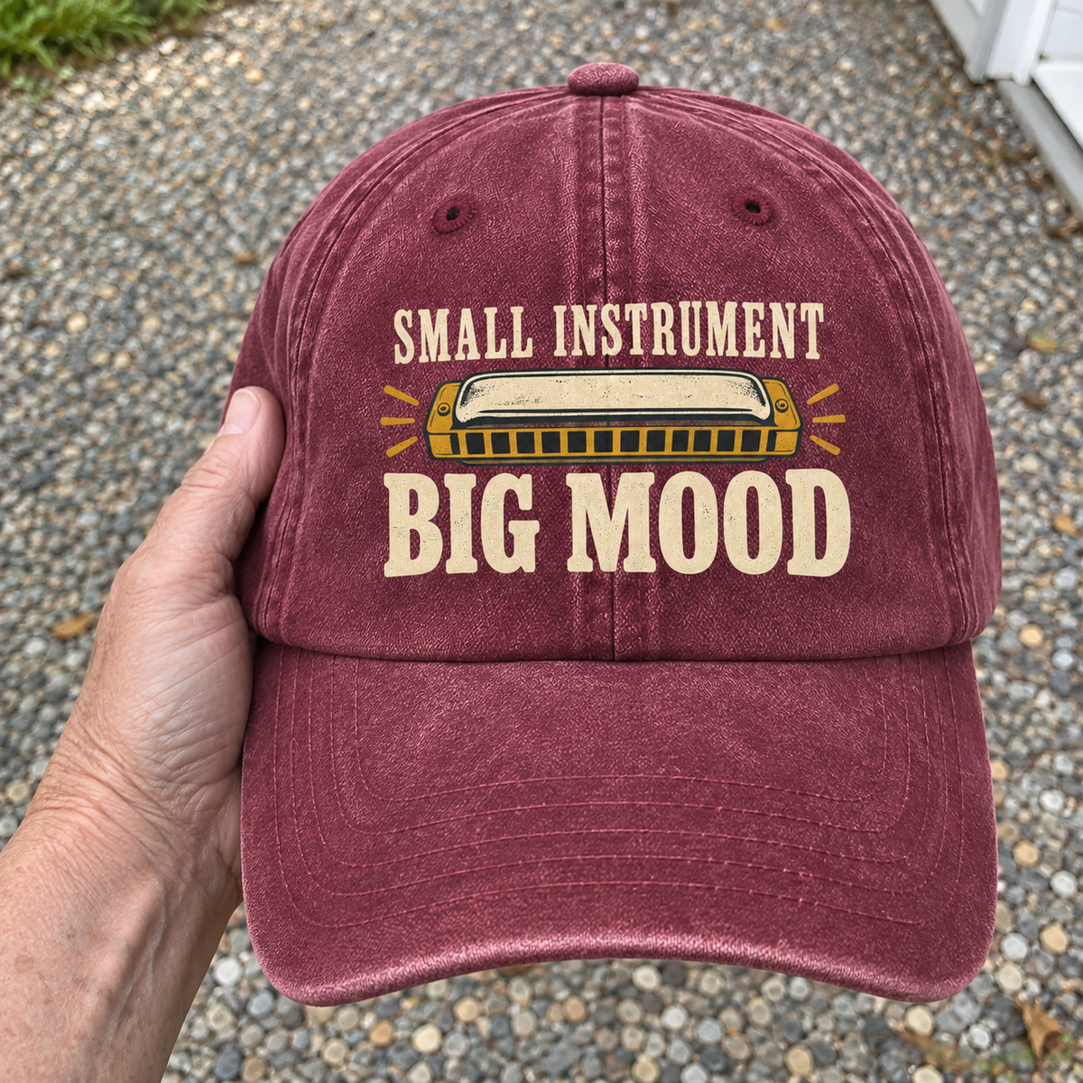 Burgundy vintage washed cap held by hand with printed Small Instrument Big Mood harmonica design