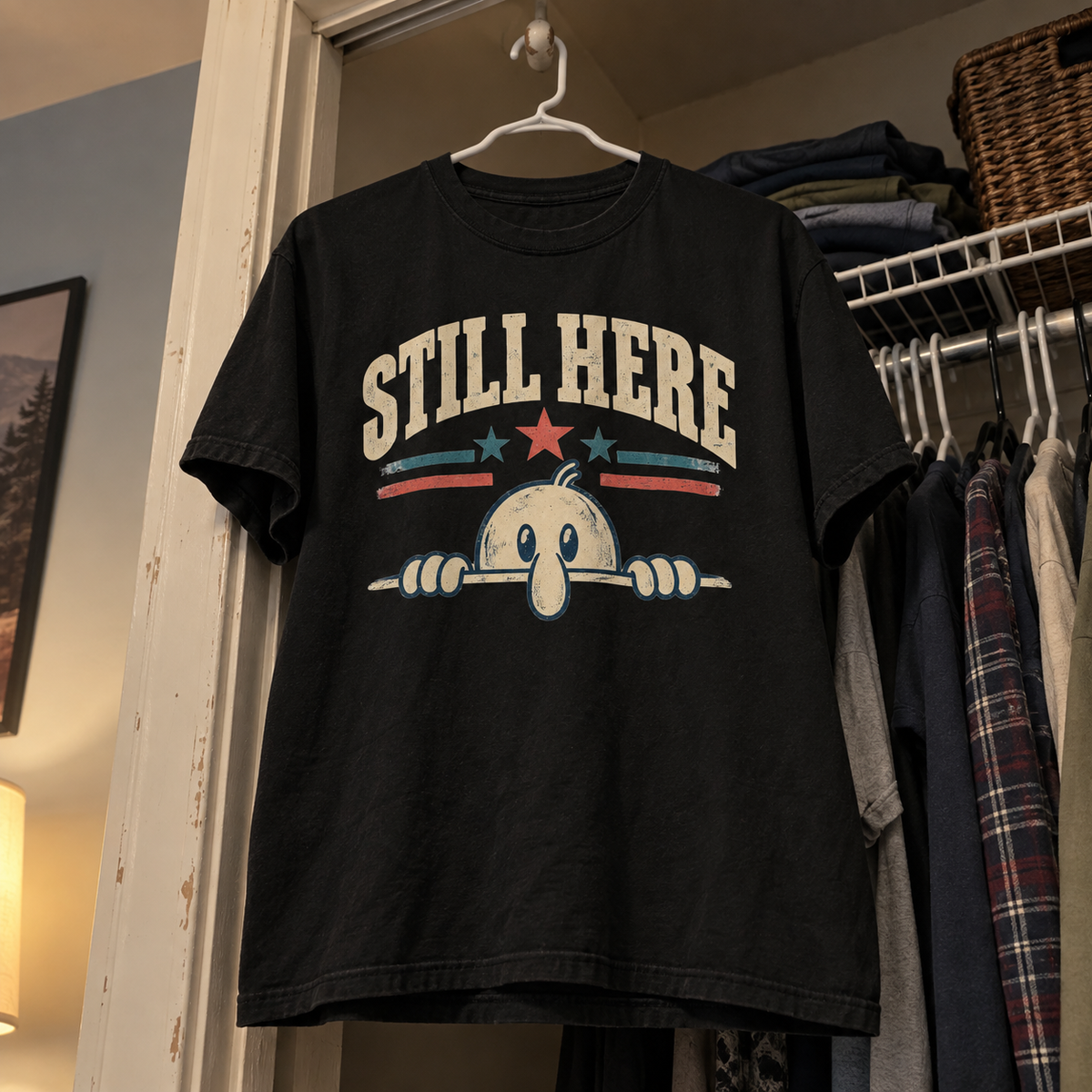 Black vintage t-shirt hanging in closet with printed Still Here slogan and peeking cartoon.