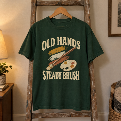 Green vintage t-shirt with printed Old Hands Steady Brush design, hanging on wooden ladder