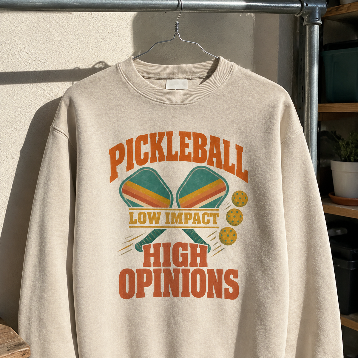 Cream vintage sweatshirt with printed pickleball paddles, balls, and Low Impact High Opinions slogan, hanging.