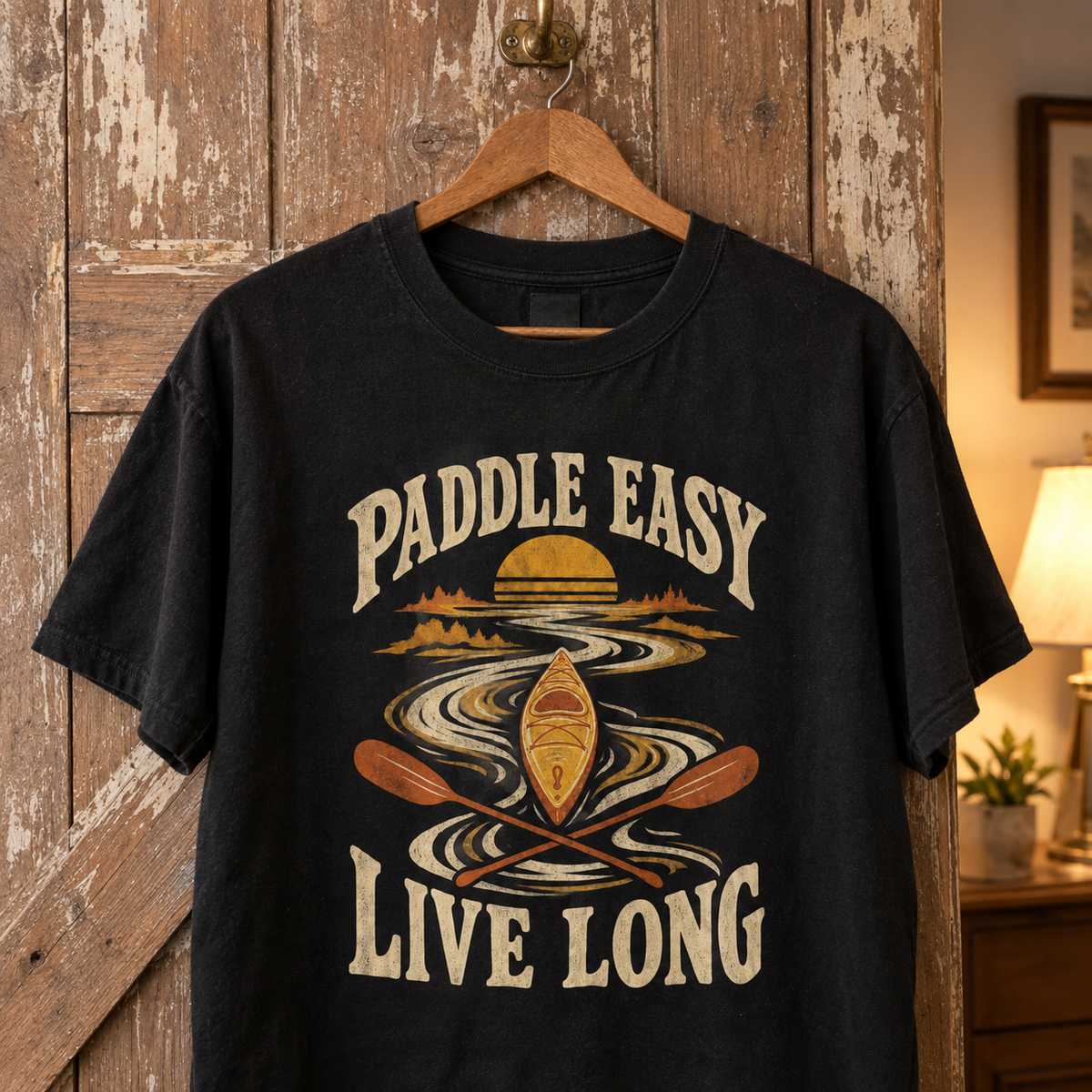 Black vintage t-shirt with printed Paddle Easy Live Long kayaking design hanging on wooden door.