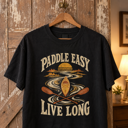 Black vintage t-shirt with printed Paddle Easy Live Long kayaking design hanging on wooden door.
