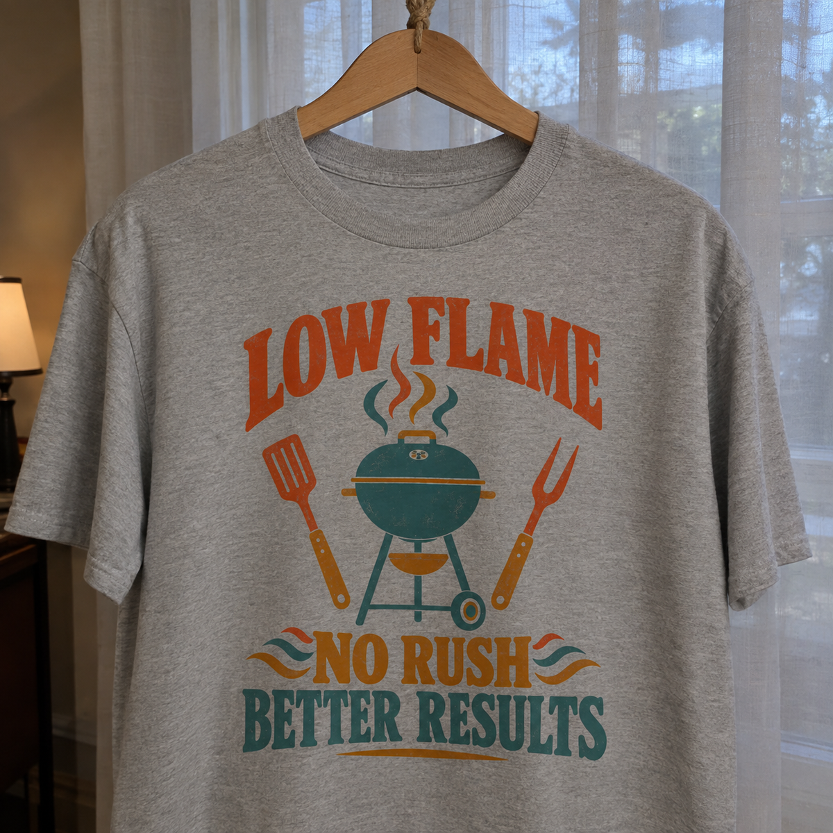 Gray vintage t-shirt hanging, printed “LOW FLAME NO RUSH BETTER RESULTS” with barbecue grill graphic.