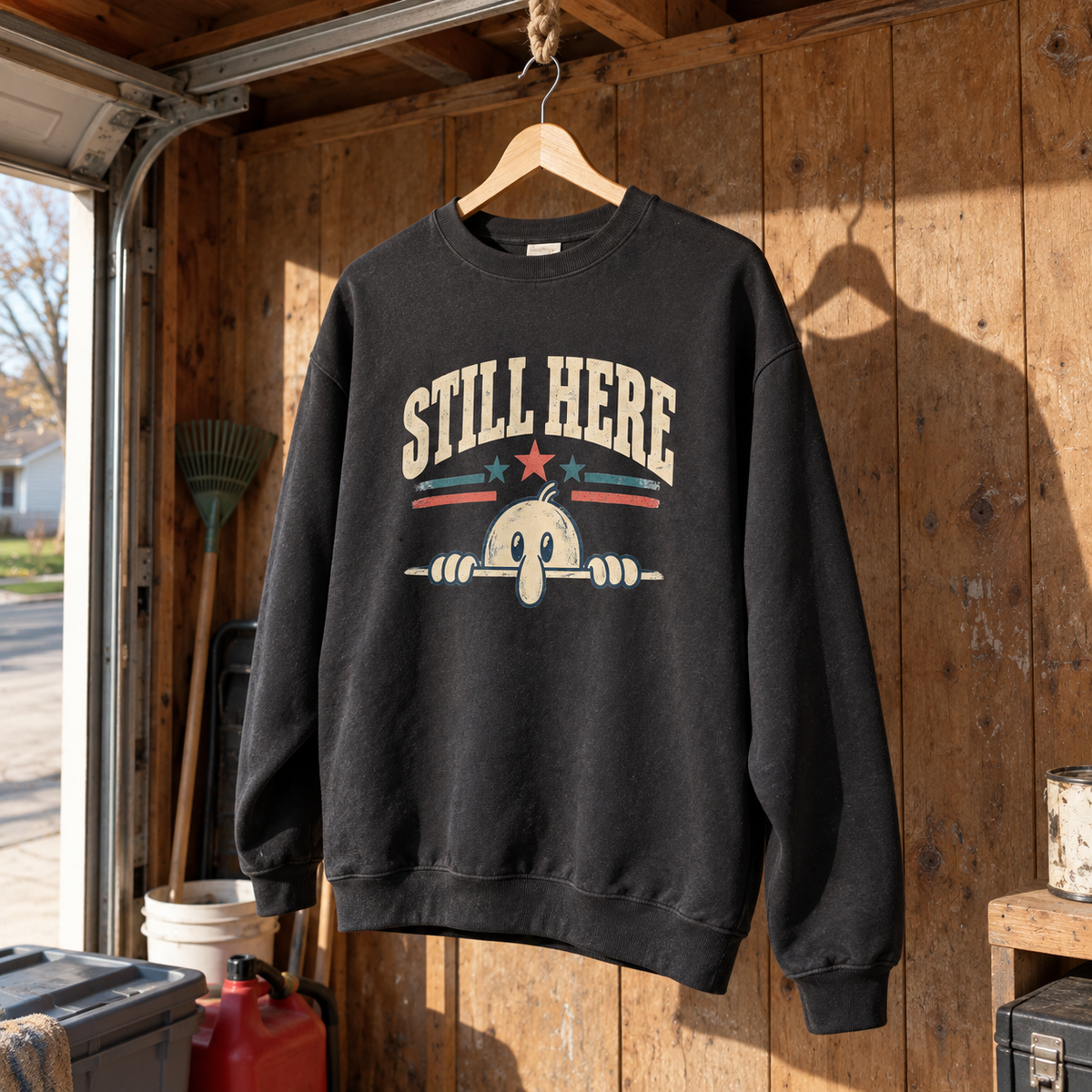 Black vintage sweatshirt hanging against wooden wall with printed Still Here cartoon graphic.