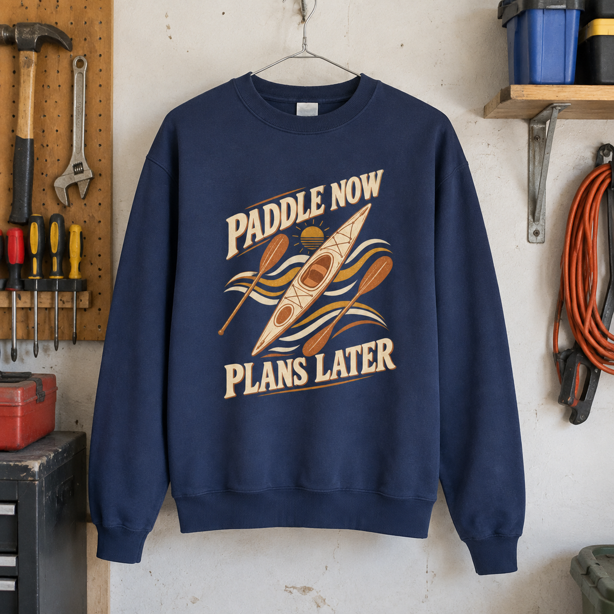 Navy vintage sweatshirt with printed “Paddle Now. Plans Later” kayak design, hanging in workshop