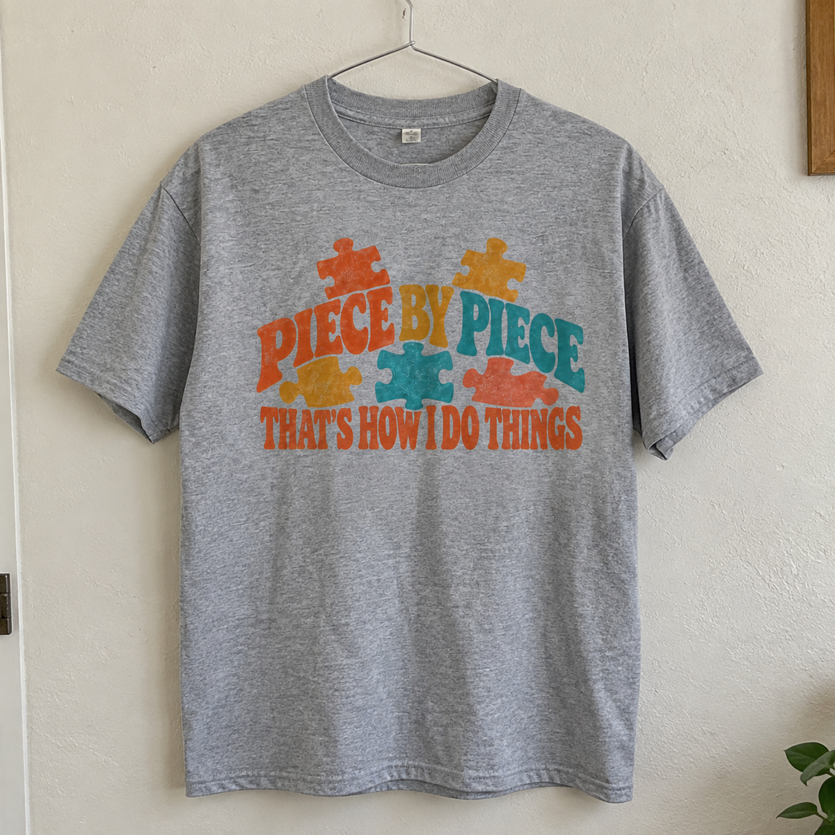 Grey vintage t-shirt hanging with colorful printed Piece By Piece slogan and puzzle designs