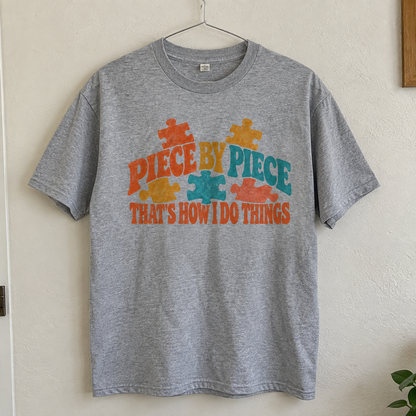 Grey vintage t-shirt hanging with colorful printed Piece By Piece slogan and puzzle designs