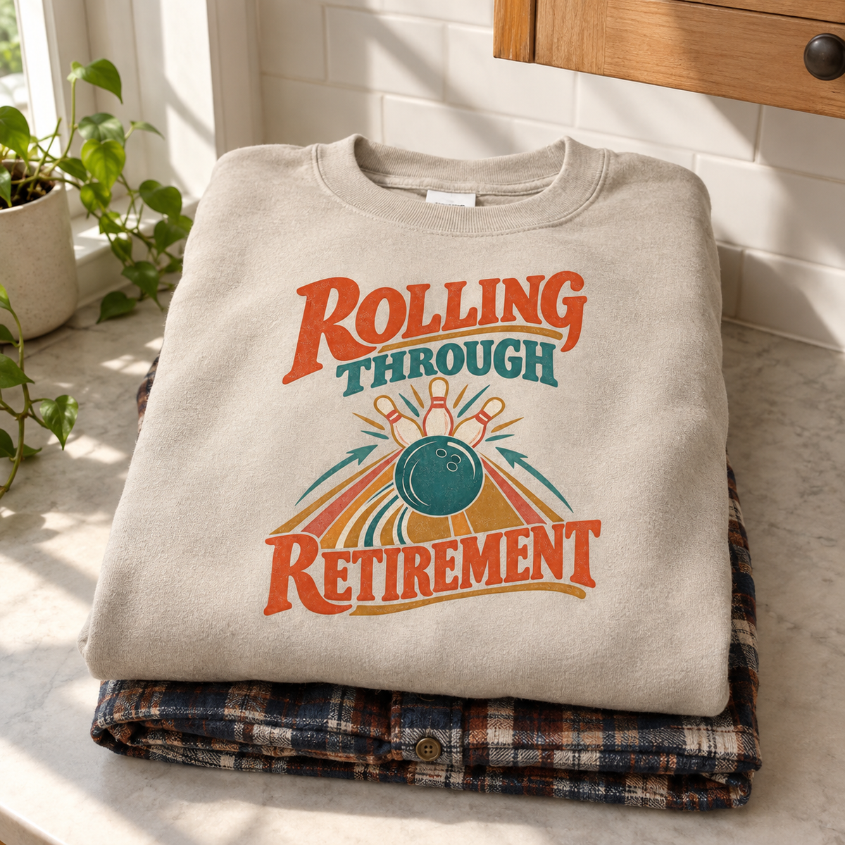 Cream vintage sweatshirt with printed Rolling Through Retirement bowling design folded on plaid shirt