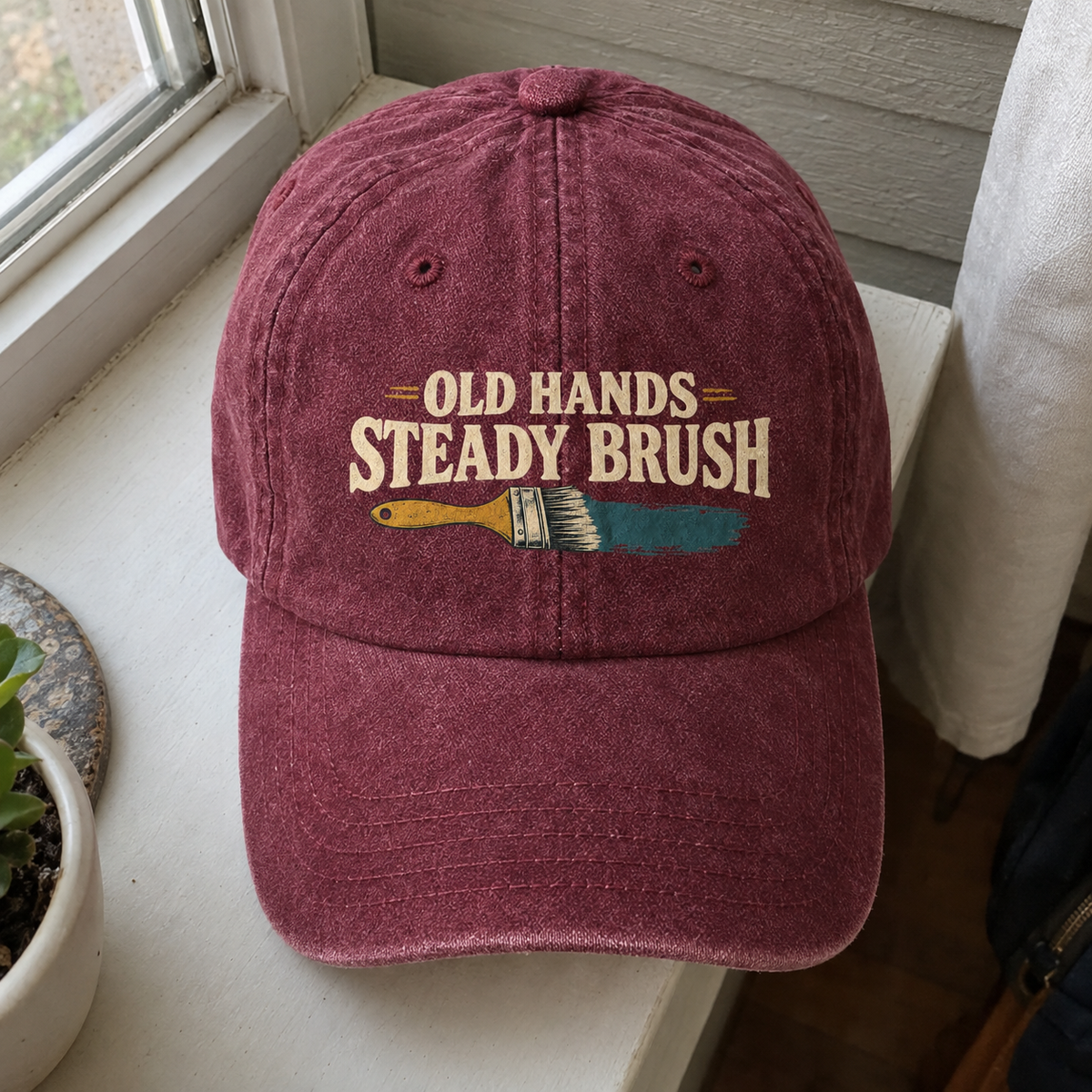 Burgundy vintage washed cap with printed “Old Hands = Steady Brush” slogan and paintbrush graphic.
