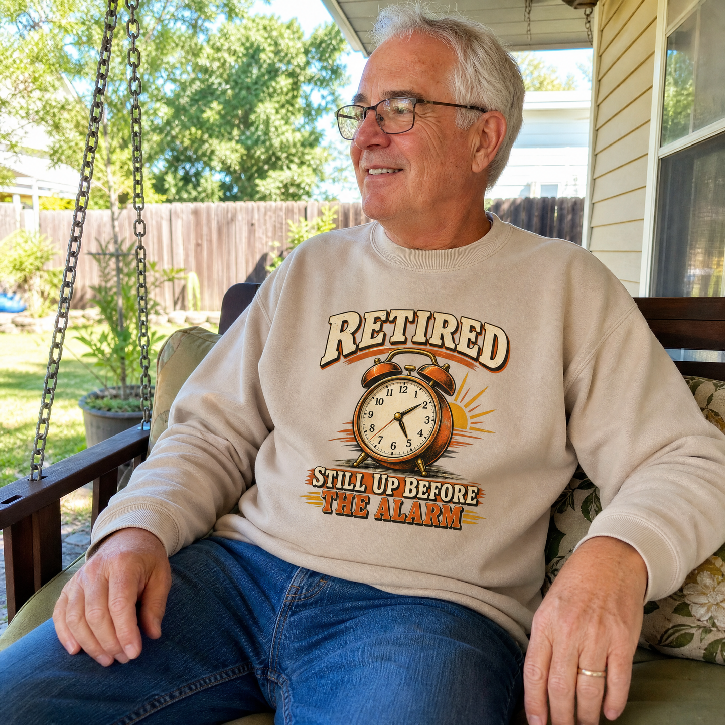 Senior man wearing vintage sweatshirt printed with Retired Still Up Before The Alarm, seated outdoors.