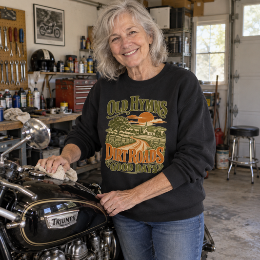 Senior woman beside motorcycle wears black vintage sweatshirt printed Old Hymns Dirt Roads Good Days.