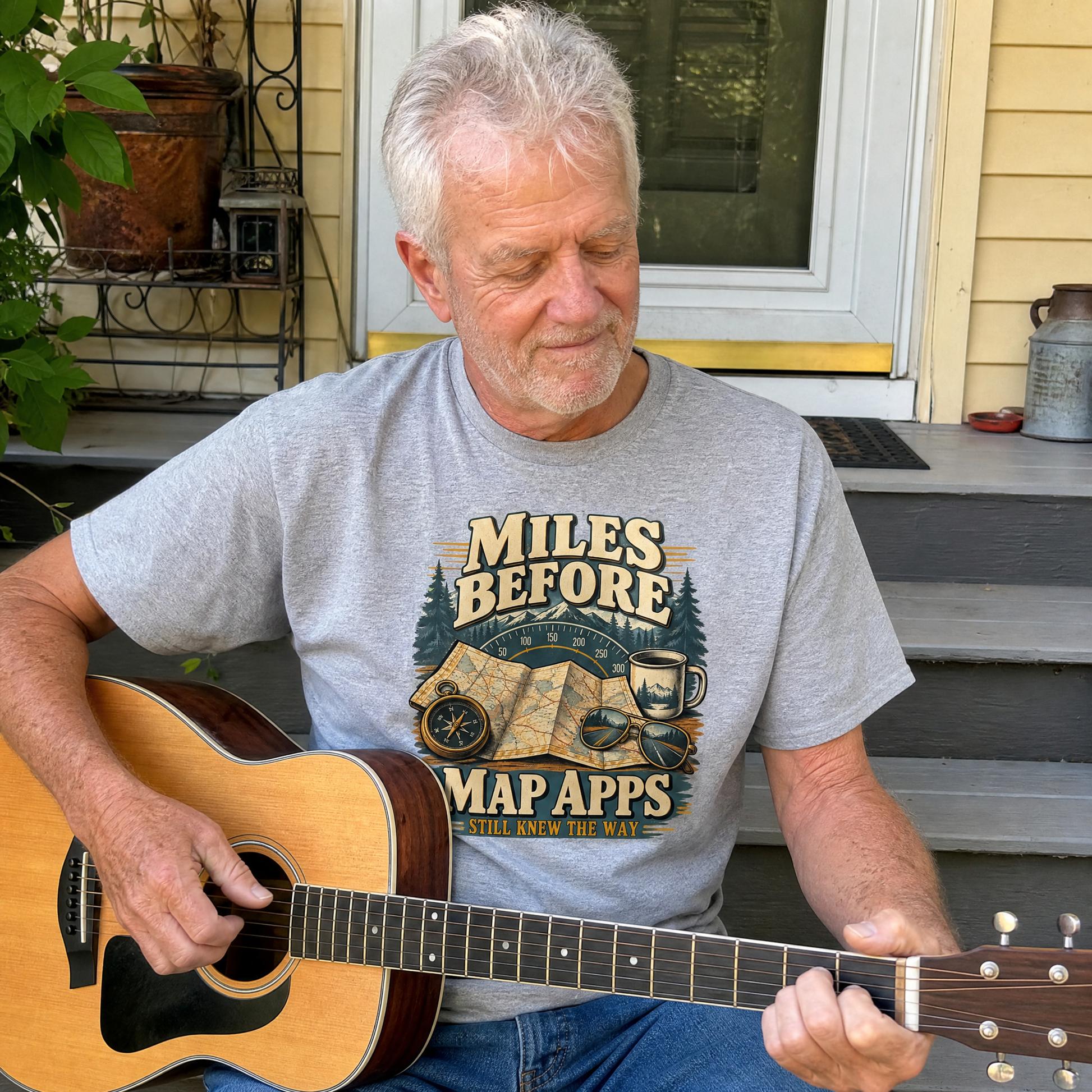 Senior man wearing gray vintage t-shirt with printed Miles Before Map Apps slogan, playing guitar.
