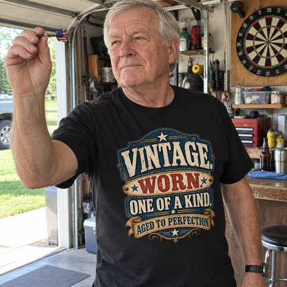 Senior man holding a dart in a garage wears black vintage t-shirt with printed slogan.