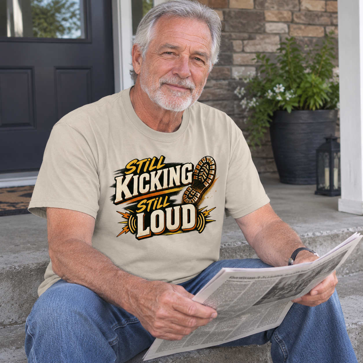 Senior man wears beige vintage t-shirt printed with Still Kicking Still Loud, seated outdoors.