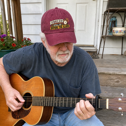 Senior man wearing burgundy vintage washed cap with printed retirement slogan, playing guitar outdoors